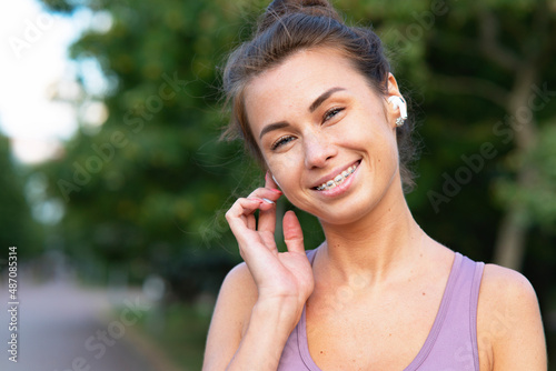 Portrait of beautiful, happy young woman talking through bluetooth headphones while at sports jog. A girl laughs, listens to music with, bluetooth wireless headphones. Funny mask with braces on teeth