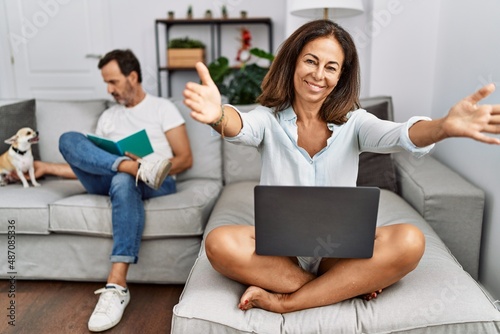 Hispanic middle age couple at home, woman using laptop looking at the camera smiling with open arms for hug. cheerful expression embracing happiness.