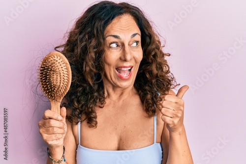 Middle age hispanic woman using comb pointing thumb up to the side smiling happy with open mouth