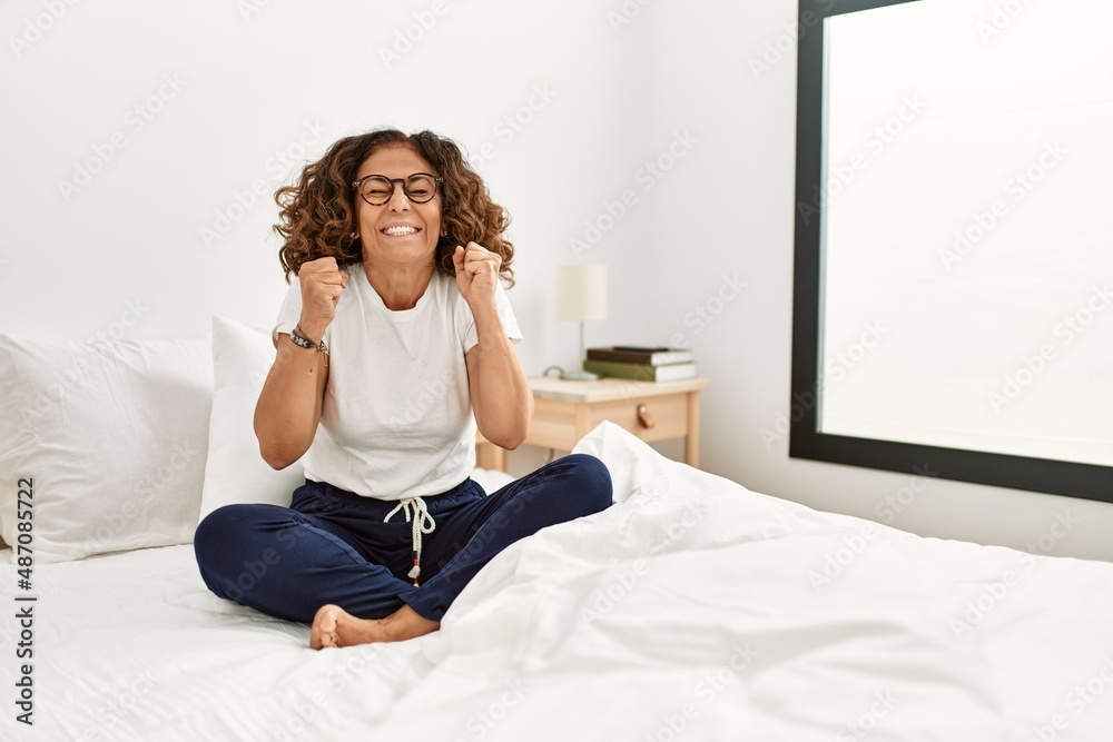 Middle age hispanic woman sitting on the bed at home excited for success with arms raised and eyes closed celebrating victory smiling. winner concept. Middle age hispanic woman sitting on the bed at home excited for success with arms raised and eyes closed celebrating victory smiling. winner concept.