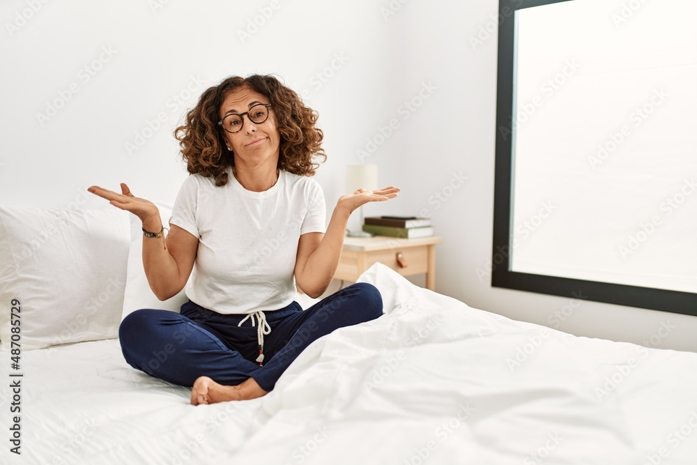 Middle age hispanic woman sitting on the bed at home clueless and confused expression with arms and hands raised. doubt concept. Middle age hispanic woman sitting on the bed at home clueless and confused expression with arms and hands raised. doubt concept.