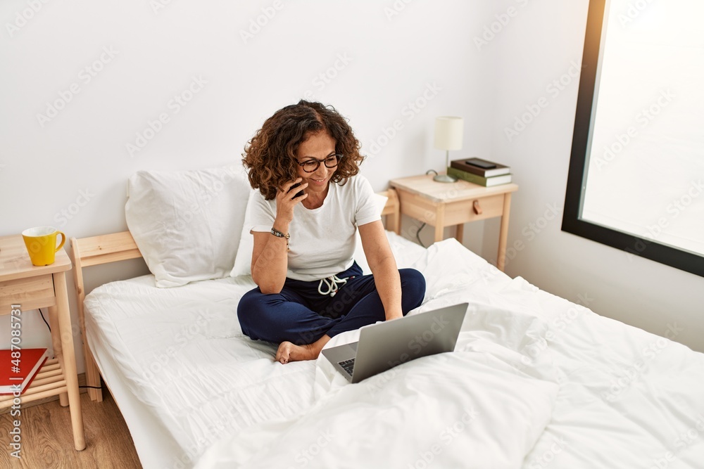 Middle age hispanic woman using laptop talking on the smartphone at bedroom