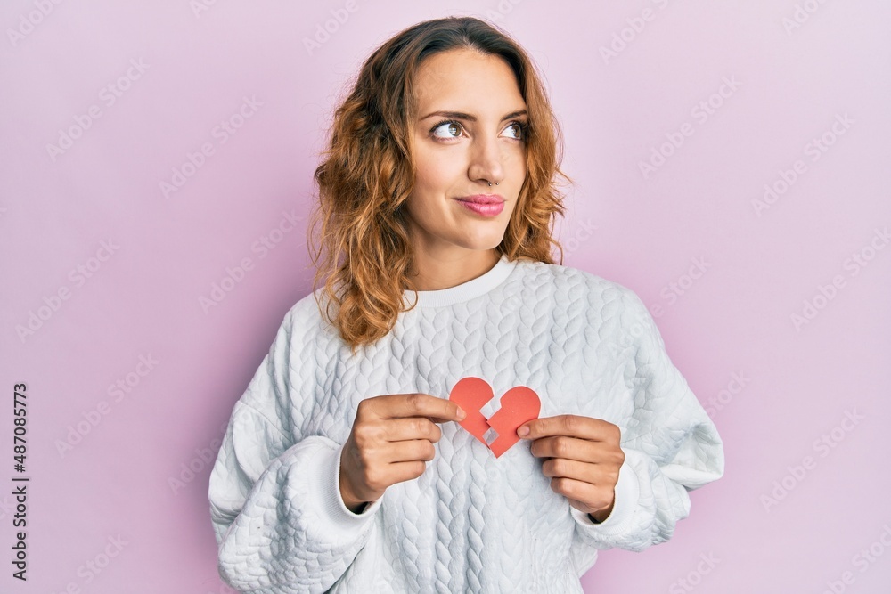 Young caucasian woman holding broken heart paper shape smiling looking to the side and staring away thinking. Young caucasian woman holding broken heart paper shape smiling looking to the side and staring away thinking.