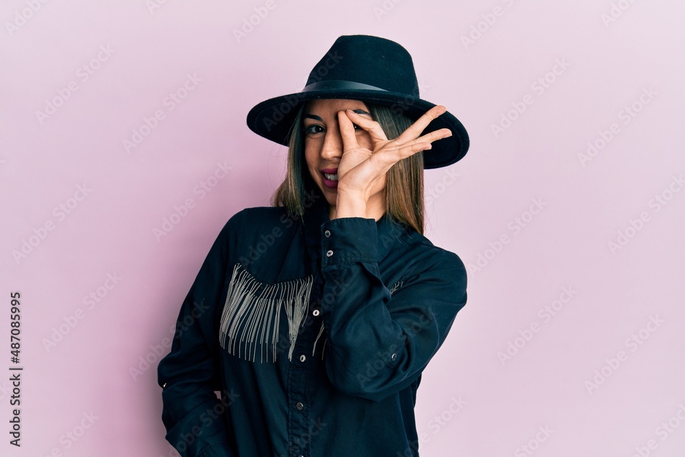 Young hispanic woman wearing cowgirl style smiling happy doing ok sign with hand on eye looking through fingers Young hispanic woman wearing cowgirl style smiling happy doing ok sign with hand on eye looking through fingers
