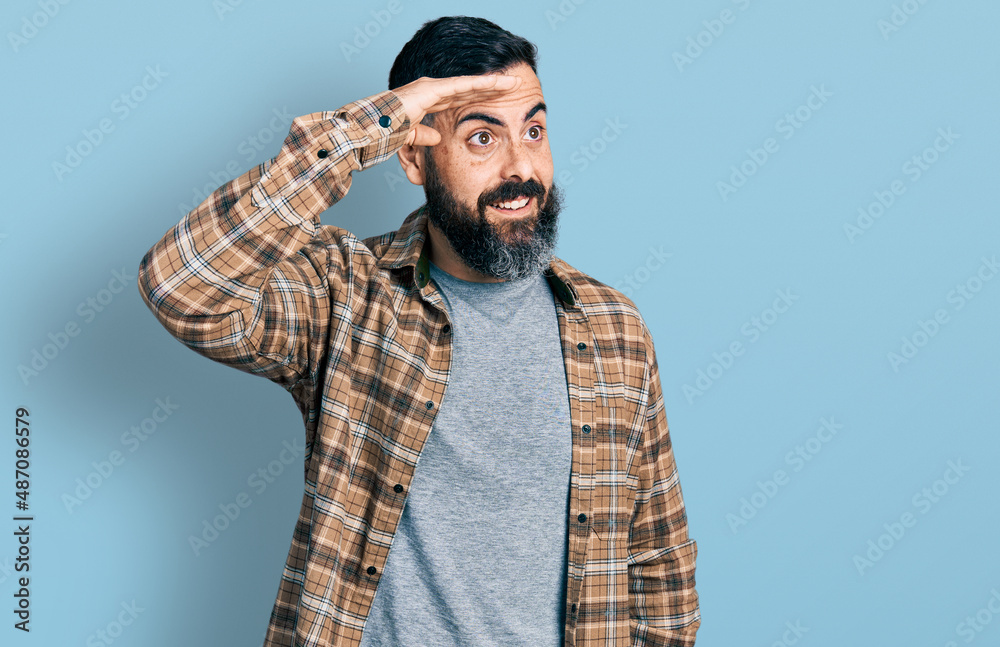 Hispanic man with beard wearing casual shirt very happy and smiling looking far away with hand over head. searching concept. Hispanic man with beard wearing casual shirt very happy and smiling looking far away with hand over head. searching concept.