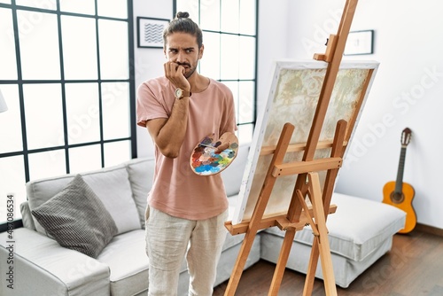 Young hispanic man with beard painting on canvas at home looking stressed and nervous with hands on mouth biting nails. anxiety problem.