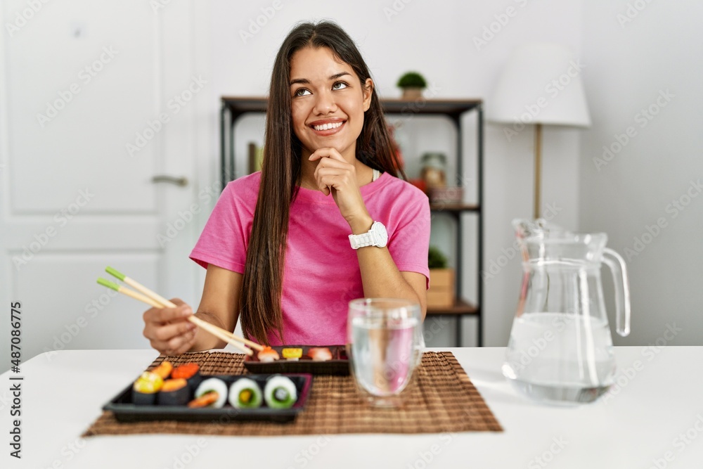 Young brunette woman eating sushi using chopsticks with hand on chin thinking about question, pensive expression. smiling and thoughtful face. doubt concept. Young brunette woman eating sushi using chopsticks with hand on chin thinking about question, pensive expression. smiling and thoughtful face. doubt concept.