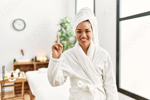 Young brunette woman wearing towel and bathrobe standing at beauty center showing and pointing up with finger number one while smiling confident and happy.