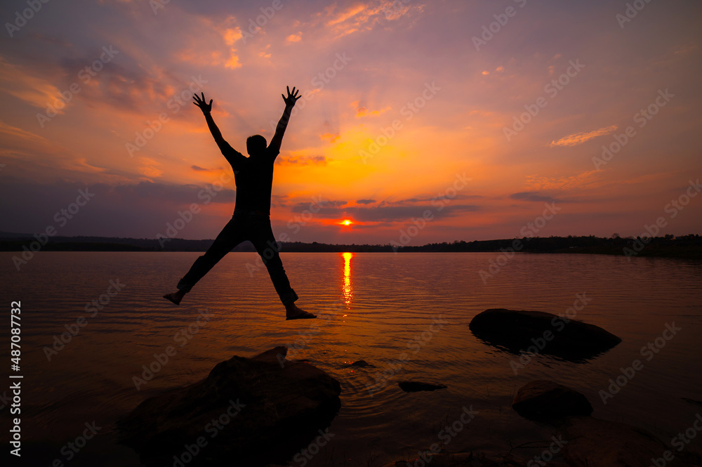 Silhouette man with backpack jumping over rocks near ocean in sunset