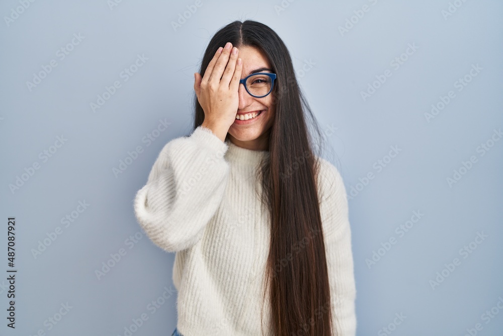 Young hispanic woman wearing casual sweater over blue background covering one eye with hand, confident smile on face and surprise emotion.