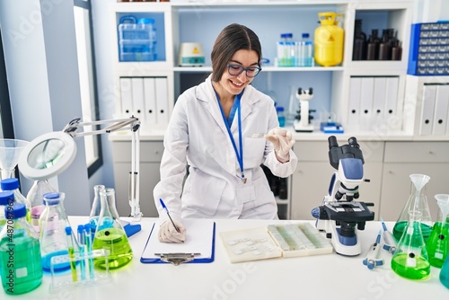 Young hispanic woman wearing scientist uniform writing on clipboard holding sample at laboratory