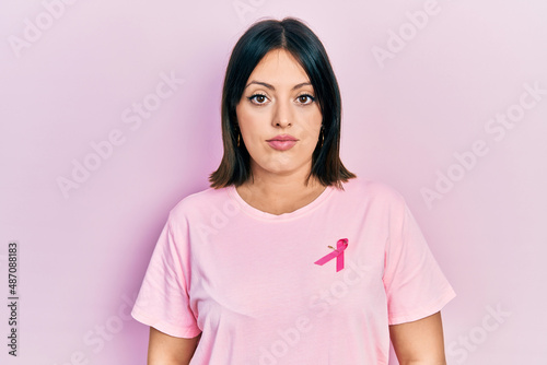 Young hispanic woman wearing pink cancer ribbon on t shirt relaxed with serious expression on face. simple and natural looking at the camera.