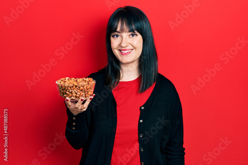 Young hispanic woman holding peanuts looking positive and happy standing and smiling with a confident smile showing teeth