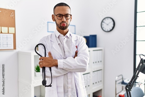 African american doctor man holding stethoscope at the clinic puffing cheeks with funny face. mouth inflated with air, catching air.