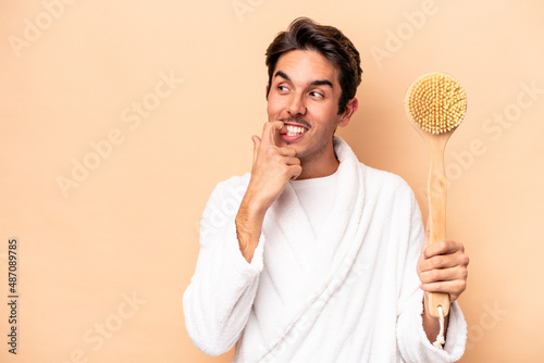 Young caucasian man wearing a bathrobe holding a back scratcher isolated on beige background relaxed thinking about something looking at a copy space.