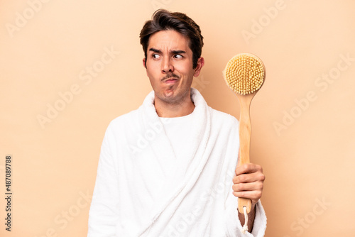 Young caucasian man wearing a bathrobe holding a back scratcher isolated on beige background confused, feels doubtful and unsure.