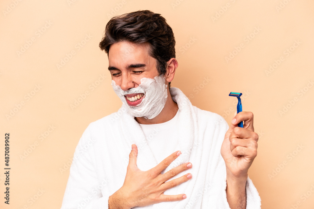 Young caucasian man shaving his beard isolated on beige background laughing and having fun.