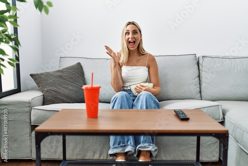 Young blonde woman eating popcorn sitting on the sofa celebrating victory with happy smile and winner expression with raised hands