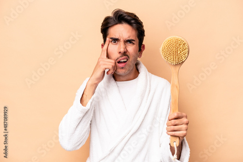Young caucasian man wearing a bathrobe holding a back scratcher isolated on beige background showing a disappointment gesture with forefinger.