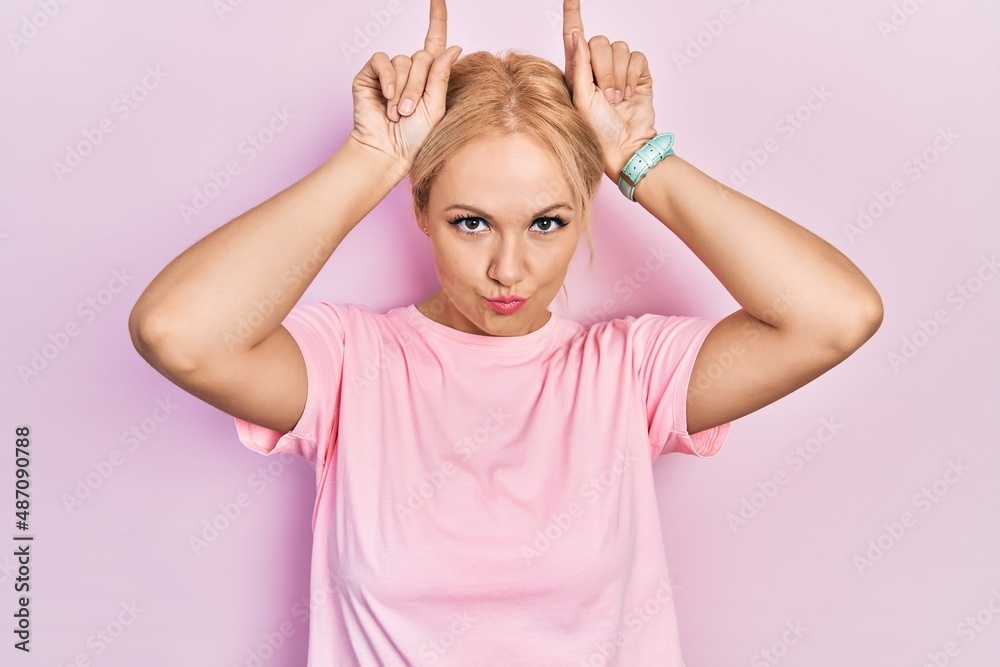 Young blonde woman wearing casual pink t shirt doing funny gesture with finger over head as bull horns Young blonde woman wearing casual pink t shirt doing funny gesture with finger over head as bull horns