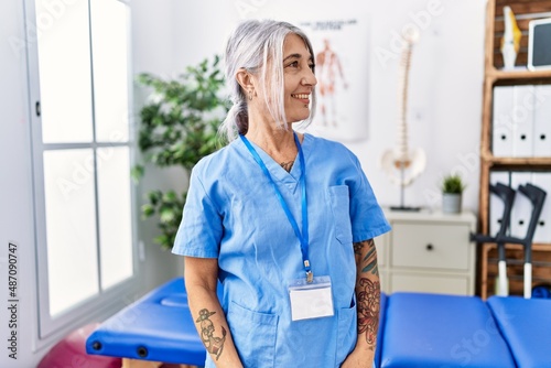 Middle age grey-haired woman wearing physiotherapist uniform at medical clinic looking away to side with smile on face  natural expression. laughing confident.