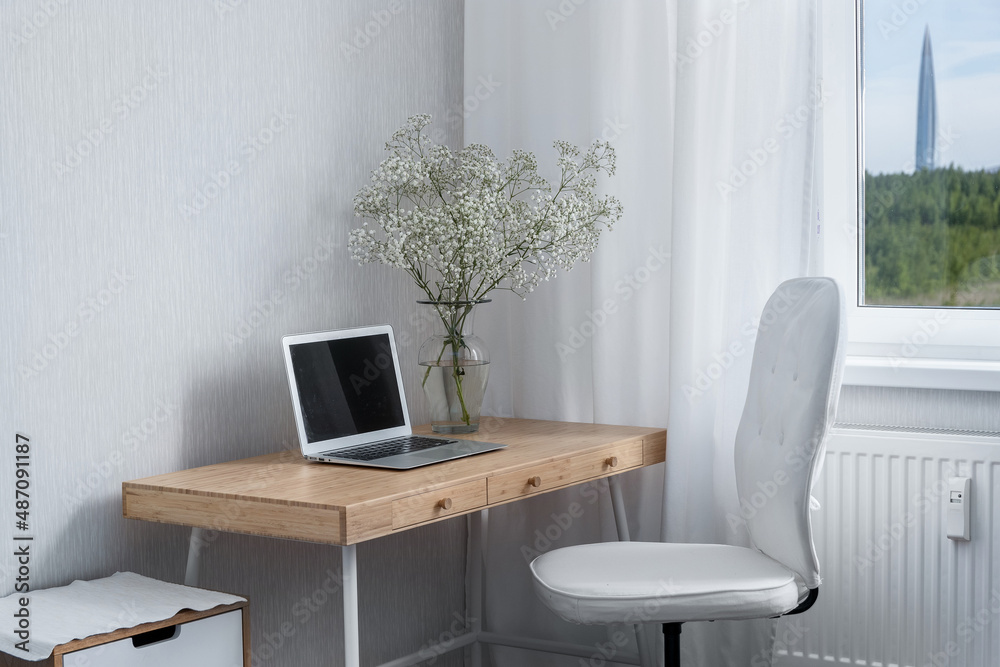 Fototapeta premium Work desk with a laptop on the table and a vase of flowers, an armchair and a view of a skyscraper in a field from the window