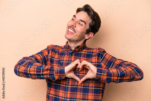 Young caucasian man isolated on beige background smiling and showing a heart shape with hands.