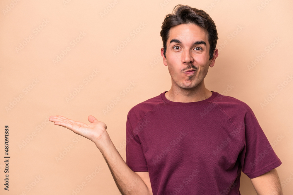 Young caucasian man isolated on beige background showing a copy space on a palm and holding another hand on waist. Young caucasian man isolated on beige background showing a copy space on a palm and holding another hand on waist.
