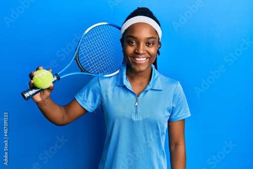 African american woman with braided hair playing tennis holding racket and ball looking positive and happy standing and smiling with a confident smile showing teeth © Krakenimages.com