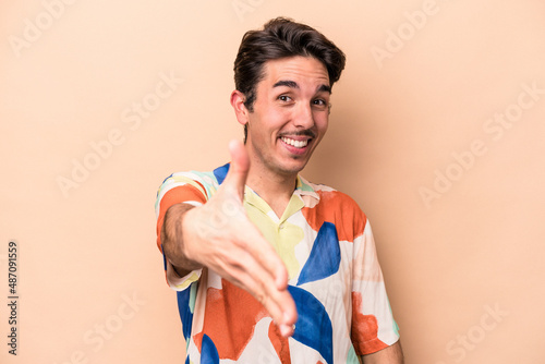 Young caucasian man isolated on beige background stretching hand at camera in greeting gesture.