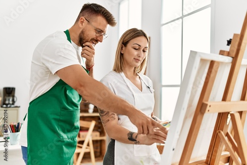 Young caucasian couple with serious expression drawing at art studio. Man standing and looking draw canvas.