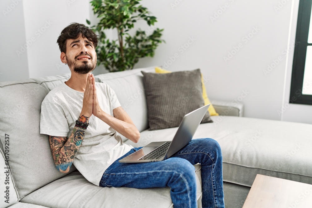 Hispanic man with beard sitting on the sofa begging and praying with hands together with hope expression on face very emotional and worried. begging. Hispanic man with beard sitting on the sofa begging and praying with hands together with hope expression on face very emotional and worried. begging.
