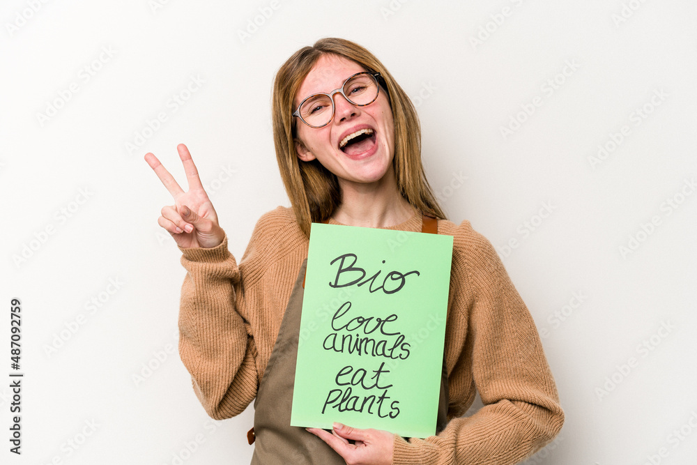 Young gardener woman holding a bio placard isolated on white background joyful and carefree showing a peace symbol with fingers. Young gardener woman holding a bio placard isolated on white background joyful and carefree showing a peace symbol with fingers.