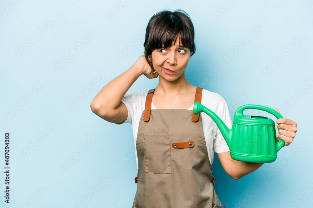 Young hispanic gardener woman holding a watering can isolated on blue background touching back of head, thinking and making a choice. Young hispanic gardener woman holding a watering can isolated on blue background touching back of head, thinking and making a choice.