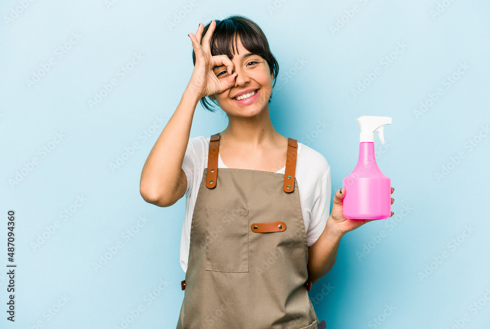 Young hispanic gardener woman holding a hand sprayer isolated on blue background excited keeping ok gesture on eye. Young hispanic gardener woman holding a hand sprayer isolated on blue background excited keeping ok gesture on eye.
