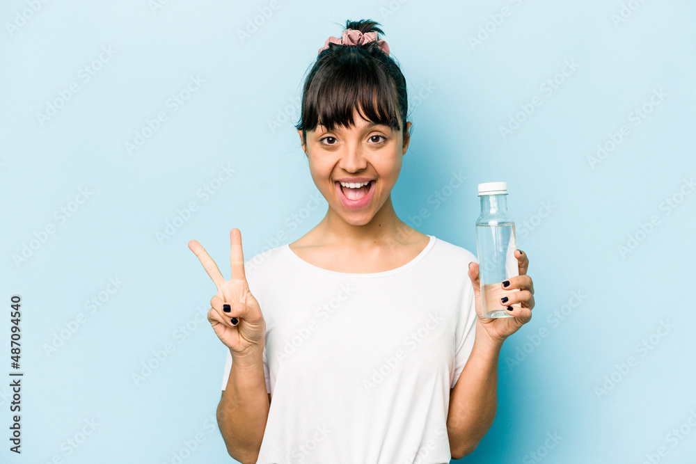 Young hispanic woman holding a bottle of water isolated on blue background joyful and carefree showing a peace symbol with fingers. Young hispanic woman holding a bottle of water isolated on blue background joyful and carefree showing a peace symbol with fingers.