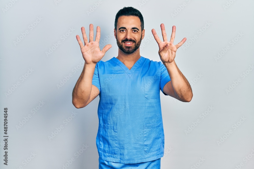 Handsome hispanic man with beard wearing blue male nurse uniform showing and pointing up with fingers number nine while smiling confident and happy. Handsome hispanic man with beard wearing blue male nurse uniform showing and pointing up with fingers number nine while smiling confident and happy.