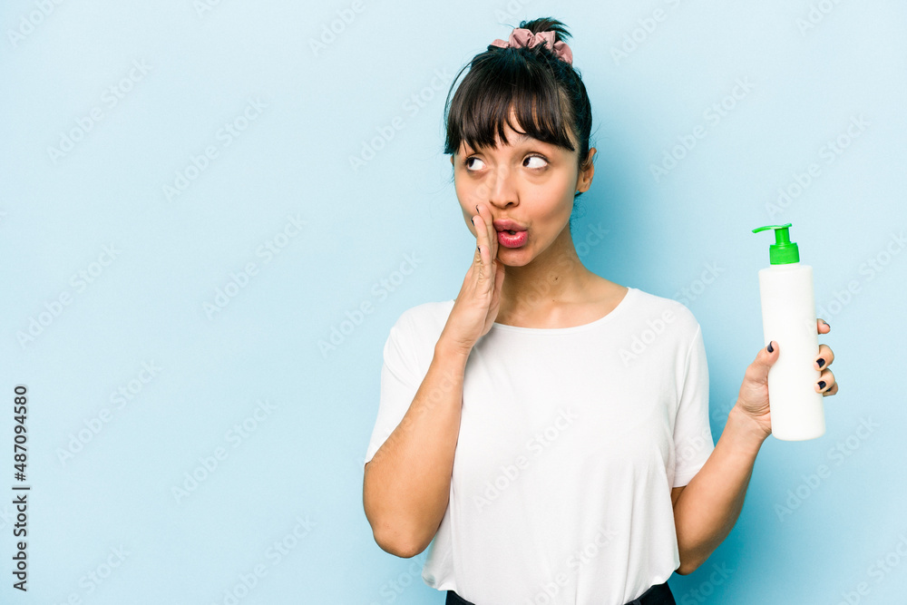 Young hispanic woman holding a body lotion isolated on blue background is saying a secret hot braking news and looking aside Young hispanic woman holding a body lotion isolated on blue background is saying a secret hot braking news and looking aside