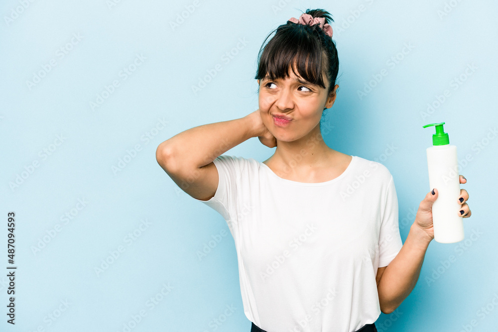 Young hispanic woman holding a body lotion isolated on blue background touching back of head, thinking and making a choice. Young hispanic woman holding a body lotion isolated on blue background touching back of head, thinking and making a choice.