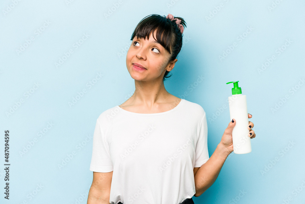 Young hispanic woman holding a body lotion isolated on blue background dreaming of achieving goals and purposes Young hispanic woman holding a body lotion isolated on blue background dreaming of achieving goals and purposes
