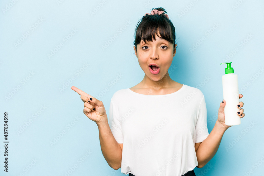 Young hispanic woman holding a body lotion isolated on blue background pointing to the side Young hispanic woman holding a body lotion isolated on blue background pointing to the side
