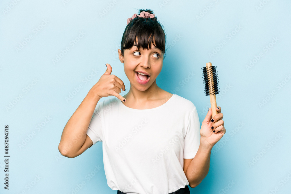 Young hispanic woman holding a brush hair isolated on blue background showing a mobile phone call gesture with fingers.