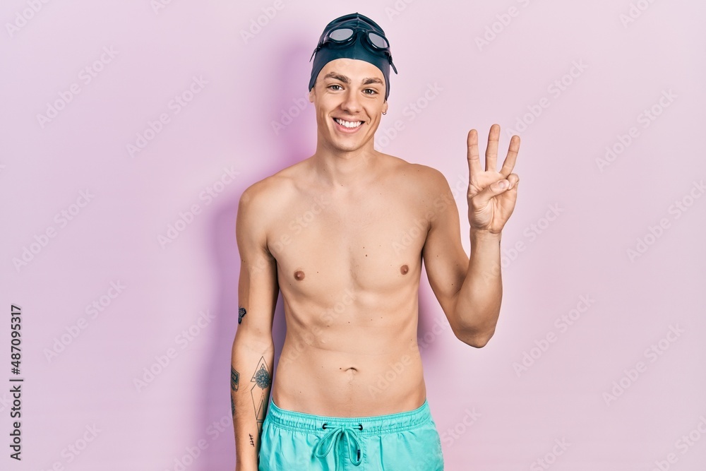 Young hispanic man wearing swimwear and swimmer glasses showing and pointing up with fingers number three while smiling confident and happy.