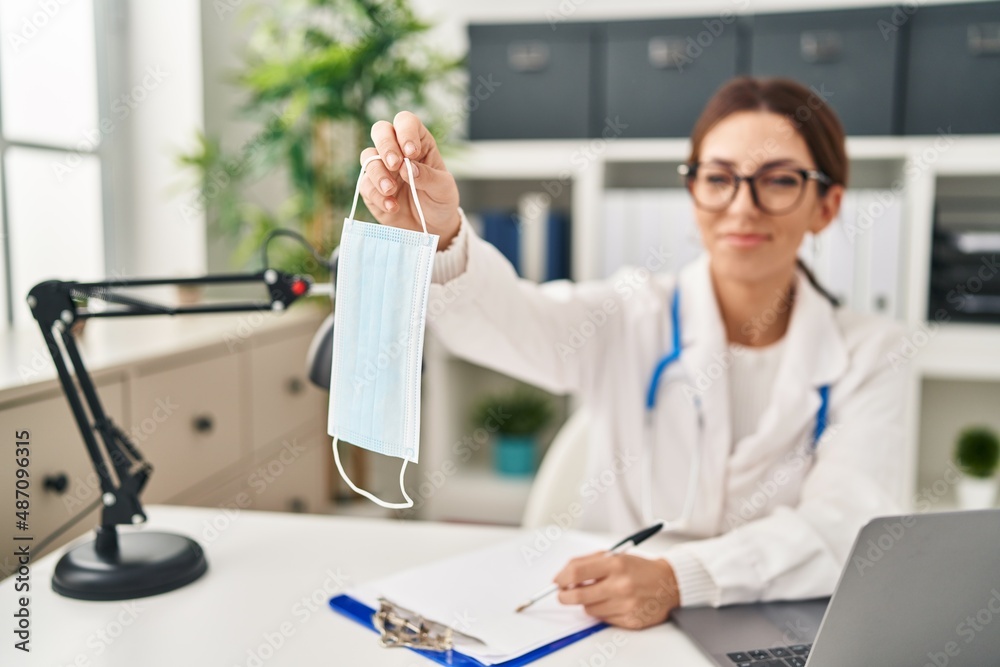 Young hispanic woman wearing doctor uniform holding medical mask at clinic Young hispanic woman wearing doctor uniform holding medical mask at clinic