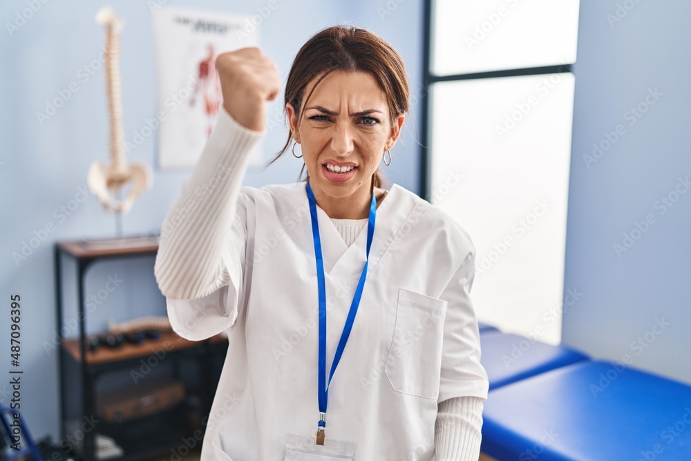 Young brunette woman working at pain recovery clinic angry and mad raising fist frustrated and furious while shouting with anger. rage and aggressive concept. Young brunette woman working at pain recovery clinic angry and mad raising fist frustrated and furious while shouting with anger. rage and aggressive concept.