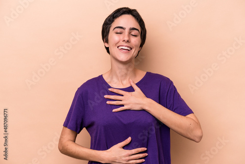 Young caucasian woman isolated on beige background laughs happily and has fun keeping hands on stomach.
