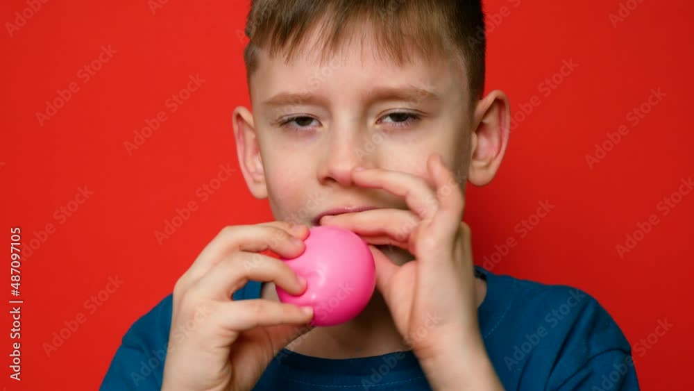 Portrait Caucasian boy 7-8 years old inflates a balloon with his mouth and looks at the camera on a red background.Sow holiday. Balloon decoration. The child inflates a pink balloon.Selective focus Portrait Caucasian boy 7-8 years old inflates a balloon with his mouth and looks at the camera on a red background.Sow holiday. Balloon decoration. The child inflates a pink balloon.Selective focus