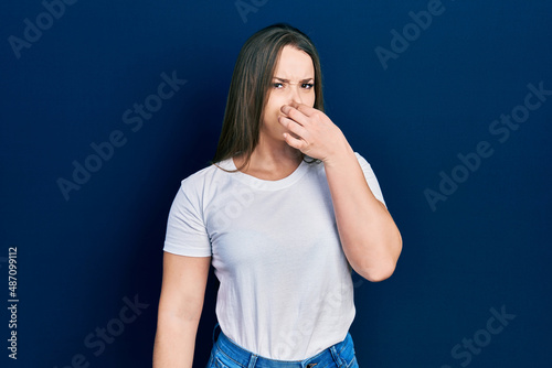 Young hispanic girl wearing casual white t shirt smelling something stinky and disgusting, intolerable smell, holding breath with fingers on nose. bad smell