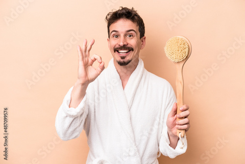 Young caucasian man holding back scratcher isolated on beige background cheerful and confident showing ok gesture.