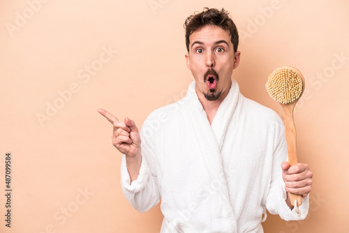 Young caucasian man holding back scratcher isolated on beige background pointing to the side